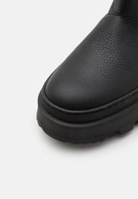 Bout de botte en cuir noir texturé avec semelle épaisse et crampons robustes, présenté en gros plan sur fond blanc.