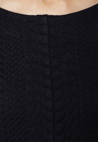 Primer plano de una prenda de punto negra, con olas texturizadas y detalles acanalados, y una costura central que corre verticalmente. Material suave.