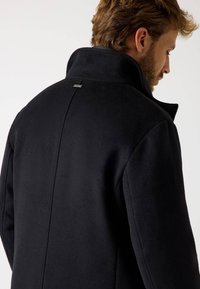 Cappotto nero con un alto colletto, realizzato in un tessuto morbido e testurizzato. Presenta un design elegante, con un sottile dettaglio del logo sul retro e linee minimaliste.