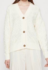Krämfärgad stickad cardigan med diamantmönster, v-ringning, fyra bruna knappar och ribbade ärmslut. Styler med veckade vita byxor.