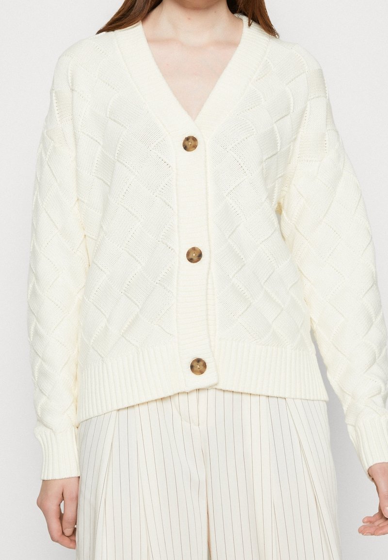 Krämfärgad stickad cardigan med diamantmönster, v-ringning, fyra bruna knappar och ribbade ärmslut. Styler med veckade vita byxor.