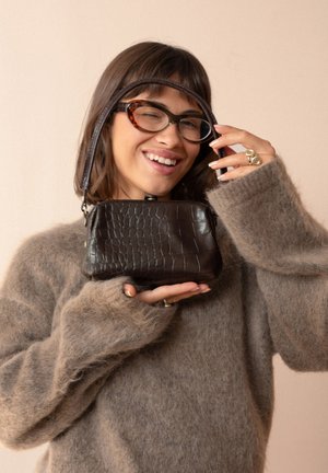 Mujer con cabello corto y castaño y gafas sonríe mientras sostiene un pequeño bolso marrón oscuro con textura, vistiendo un suéter borroso color topo.