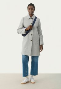 Grauer Wollmantel mit Kragen, drei Knöpfen und zwei seitlichen Taschen, kombiniert mit blauen geraden Jeans und weißen Sneakern.