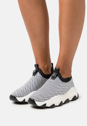 Pieds portant des baskets en maille noir et blanc à enfiler avec des semelles blanches épaisses et texturées et des languettes noires sur un fond clair.