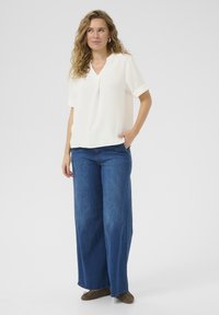 Blusa blanca de manga corta texturizada con escote en V y puños enrollados, combinada con jeans de mezclilla azul de pierna ancha y zapatos marrones.