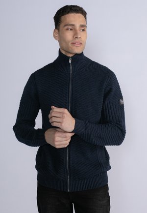 Marine blauwe ritstrui met hoge kraag, met textuur in horizontale en verticale ribbels. Bevat een logo op de linker mouw, strakke manchetten en taille.