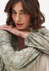 Top in pizzo floreale verde chiaro con maniche elastiche, sovrapposto a un capo beige morbido. La modella posa con le braccia incrociate, sfoggiando un trucco naturale e capelli di lunghezza spalle.