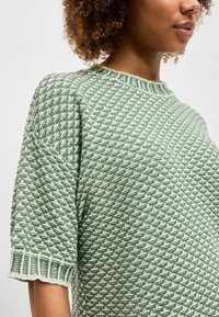 Nő világoszöld, texturált kötött rövid ujjú pulóverben, bordázott kerek nyakkivágással és mandzsettákkal, vállaktól a törzs közepéig látható.