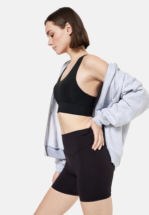 Vrouw draagt een zwarte sportbeha en short met een lichtgrijze hoodie met rits, staat met hand op heup en kijkt omlaag in profiel.