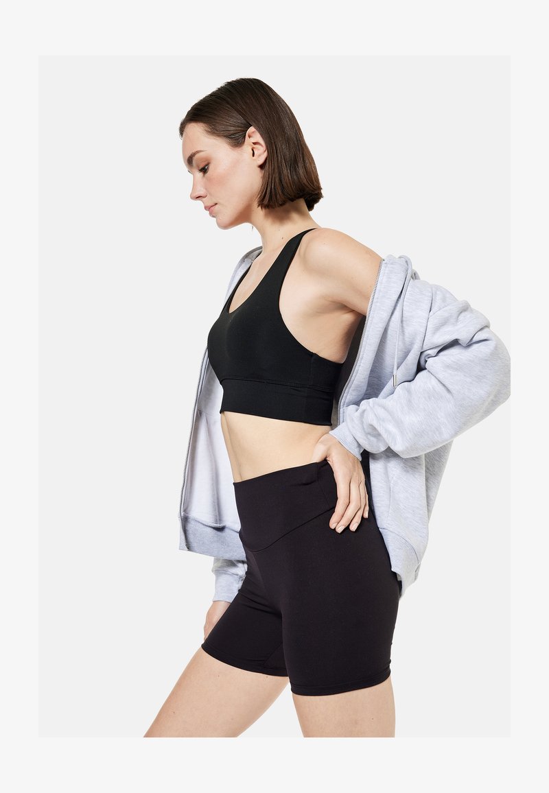 Vrouw draagt een zwarte sportbeha en short met een lichtgrijze hoodie met rits, staat met hand op heup en kijkt omlaag in profiel.