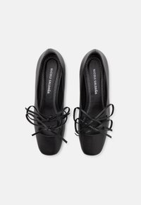 Zapatos planos de cuero negro con punta redondeada, con cordones negros prominentes atados en un lazo. Textura suave con costuras en blanco contrastantes.
