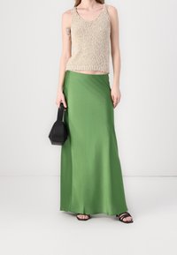 Top de punto beige sin mangas con escote redondo, combinado con una falda maxi de satén verde. La modelo sostiene un bolso geométrico negro. Sandalias negras con tiras.