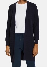 Granatowy sweter z dzianiny z otwartym przodem, ściągaczami na mankietach i fakturowanym wzorem. Nosi się go na białej koszuli w zestawie z ciemnymi spodniami.