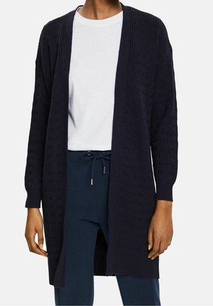 Cardigan tricoté marine avec une ouverture à l'avant, des poignets côtelés et un motif texturé. Porté sur une chemise blanche et associé à un pantalon foncé.