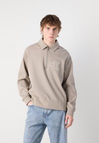 Hanorac crem cu guler tip fermoar, având un design texturat, cu un logo CK brodat pe piept și manșete cu rebrare. Asortat cu blugi din denim de culoare albastru deschis.