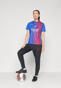 Mėlynas ir raudonas futbolo marškinėliai su trumpomis rankovėmis ir logotipu, suderinti su juodomis kelnėmis, turinčiomis raudoną Nike swoosh, stovintis ant futbolo kamuolio.