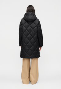 Nainen, joka pukeutunut mustaan tikattuun hupulliseen takkiin ja leveisiin beigenvärisiin housuihin, seisoo selin valkoista taustaa vasten.