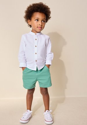 Copil mic cu păr creț, purtând o cămașă albă cu nasturi, pantaloni scurți verzi și adidași albi, stă cu mâinile în buzunare pe un fundal bej.