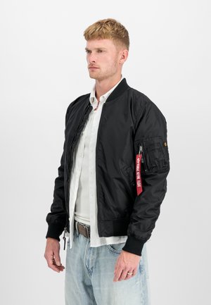 Black bomber jacket with zip closure, ribbed cuffs, and pocket on the left sleeve. Features red fabric tag. Worn over a white shirt.