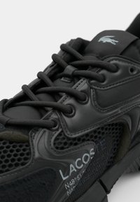 Zapatillas deportivas negras con parte superior de malla texturizada, acentos suaves y diseño de cordones. Marca destacada en el costado. Cordoneras negras mate.