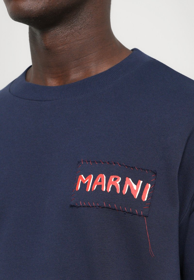Man wearing a navy blue shirt with a hand-stitched patch on the chest that reads "MARNI" in white and red letters.