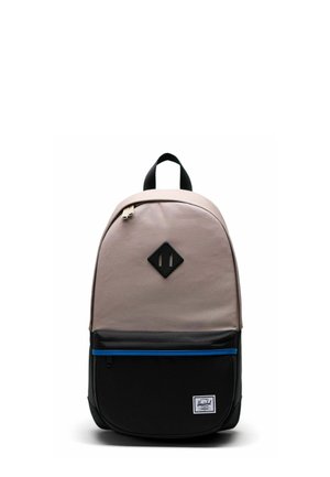 Sac à dos avec le dessus beige, poche inférieure noire avec une fermeture éclair bleue, poignée noire et écusson noir en forme de losange sur le devant, sur fond blanc.