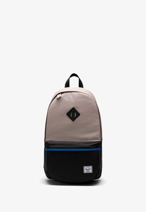 Sac à dos avec le dessus beige, poche inférieure noire avec une fermeture éclair bleue, poignée noire et écusson noir en forme de losange sur le devant, sur fond blanc.