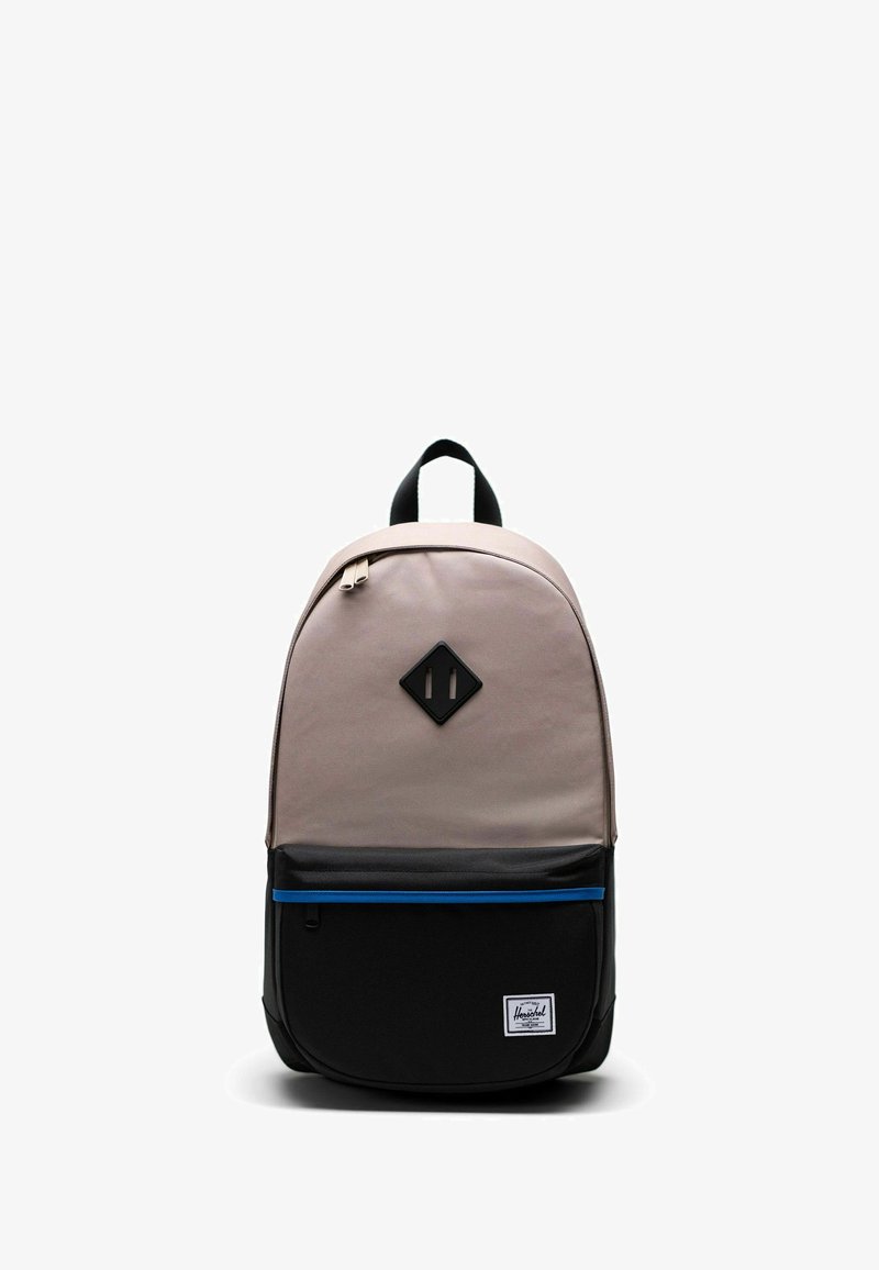 Sac à dos avec le dessus beige, poche inférieure noire avec une fermeture éclair bleue, poignée noire et écusson noir en forme de losange sur le devant, sur fond blanc.