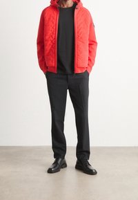 Man i en röd vadderad zip-up hoodie, svart crew neck-tröja, svarta byxor och svarta läderklackskor står på ett grått golv.