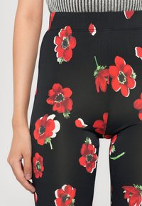 Nærbilde av en persons nedre torso iført svarte bukser med lys røde blomstermønster og grønne detaljer, kombinert med en grå ribbet topp.