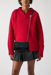 Roter Strickpullover mit V-Ausschnitt und geripptem Saum, kombiniert mit einer schwarzen gesteppten Umhängetasche mit goldenem Logo. Sichtbarer grauer karo-gefalteter Rock.