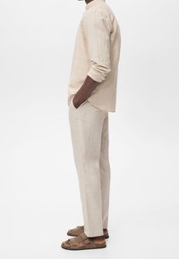 Homme debout de profil, portant une chemise beige à manches longues, un pantalon léger en lin et des sandales marron à double lanière, les mains dans les poches.