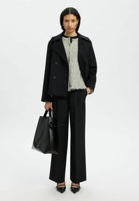 Kobieta ubrana w czarną kurtkę, szary sweter, szerokie czarne spodnie, czarne buty na szpic i trzymająca dużą czarną torebkę, stojąca na białym tle.
