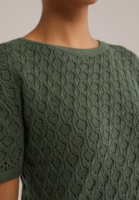 Zelený pletený svetr s texturovaným, otevřeným vzorem. Má kulatý výstřih a krátké rukávy, které předvádějí geometrický design.