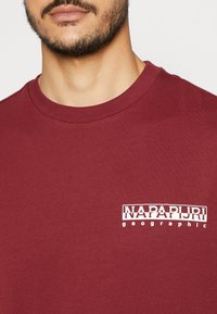 Vousatý muž oblečený v vínovém crewneck tričku s malým bílým logem "NAPAPIJRI geographic" na levé části hrudi.