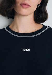 Juodas trumpas rankovės marškinėliai su apvalia iškirpte, turintys baltą "HUGO" logotipą ir kontrastingas pilkas siūlas aplink iškirptę.