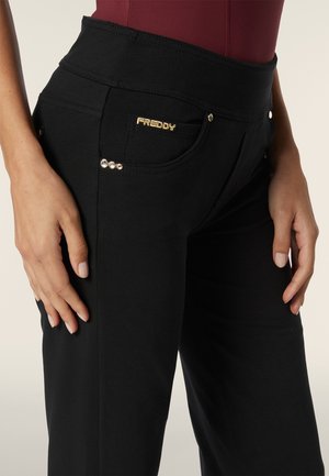Pantaloni neri elasticizzati con un alto punto vita, dotati di un logo dorato, tasche decorative e dettagli metallici lungo la tasca anteriore.