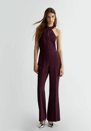 Tutina bordeaux con scollatura alta, dettaglio a cut-out e gambe svasate. Il tessuto ha una texture liscia; i tacchi alti argento completano l'outfit.