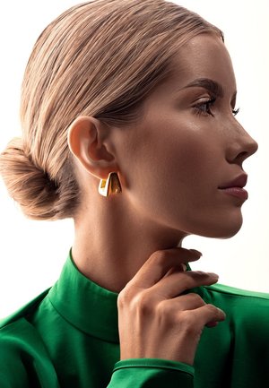 Złote geometryczne kolczyki o gładkiej fakturze, noszone przez modelkę z elegancką fryzurą, zestawione z zieloną bluzką z wysokim kołnierzem.