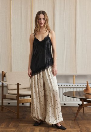 Donna in piedi all'interno con indosso una canottiera nera bordata di pizzo e pantaloni larghi color crema con pois neri, accanto a una sedia in legno e a un tavolo rotondo.