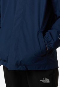 Niebieska kurtka wodoodporna z elastycznymi mankietami, kieszeniami na suwak i gładką teksturą. Posiada branding na rękawie i dole kurtki.