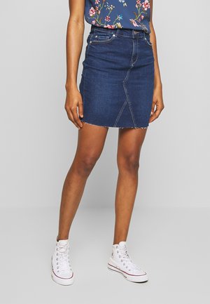 Femme portant une mini-jupe en denim bleu foncé à ourlet brut, un haut fleuri et des baskets montantes blanches, debout devant un fond uni.