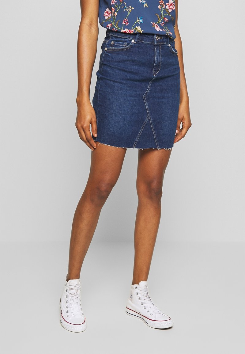 Femme portant une mini-jupe en denim bleu foncé à ourlet brut, un haut fleuri et des baskets montantes blanches, debout devant un fond uni.