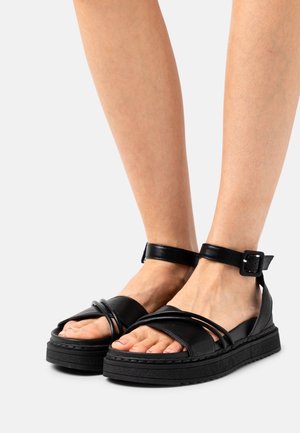 Pieds portant des sandales à brides cheville en cuir noir avec des semelles épaisses et plusieurs fines brides sur les orteils sur un fond gris clair.