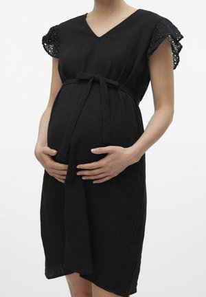 Těhotná žena v černých šatech s krajkovými rukávy, která drží břicho oběma rukama svázanýma látkovým pásem.