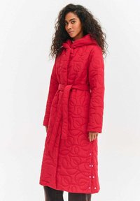 Czerwony pikowany długi płaszcz z paskiem w talii, kapturem i wzorem liści. Posiada zatrzaski po bokach dla regulowanego dopasowania i wygody.