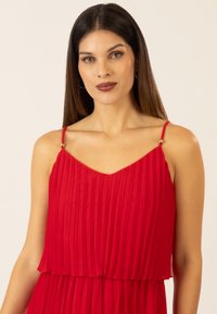 Rotes, plissiertes Kleid mit V-Ausschnitt, dünnen Trägern und goldenen Akzenten an den Schultern; leichtes Gewebe mit fließender Silhouette.