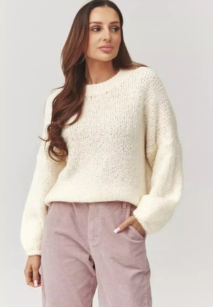 Kobieta o długich brązowych włosach, ubrana w biały dziergany sweter i jasnofioletowe spodnie sztruksowe, stojąca z jedną ręką w kieszeni.