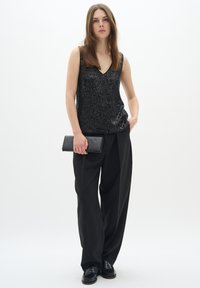 Top senza maniche nero con paillettes e scollatura a V, abbinato a pantaloni neri larghi. Il modello tiene una pochette nera con una mano.