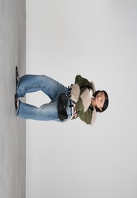 Γυναίκα που φορά πράσινο μπουφάν από γούνινη ύφανση, μπλε φαρδιά τζιν, μαύρες σαγιονάρες και κρατάει μια μαύρη τσάντα ώμου, στέκεται μπροστά σε απλό φόντο.